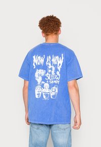 Modré bavlněné tričko s velkým bílým grafickým potiskem s textem "TEĎ JE TEĎ" a ilustracemi rostlin v květináčích na zadní straně.