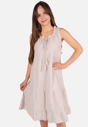 Jeune femme aux longs cheveux bruns portant une robe beige sans manches à volants, longueur genou, avec un lien à pompons au niveau du décolleté.
