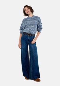 Woman wearing a blue patterned knit sweater, high-waisted wide-leg denim jeans, brown belt, and brown shoes, standing with one hand in pocket.