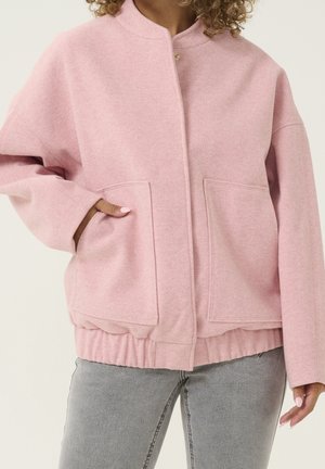 Kvinde iført en lyserød, lynlåstrøje med store forlommer og elastisk kant, kombineret med lysegrå jeans.