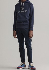 Felpa navy con logo bianco "GANT", pantaloni del sudore grigi e sneakers multicolori (blu, rosso, giallo) con suola testurizzata.