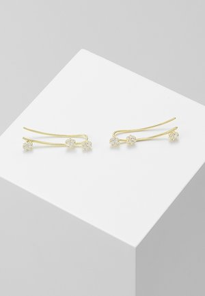 Boucles d'oreilles escaladeurs en or avec trois petites fleurs en cristal clair sur chaque, présentées sur une surface géométrique blanche.