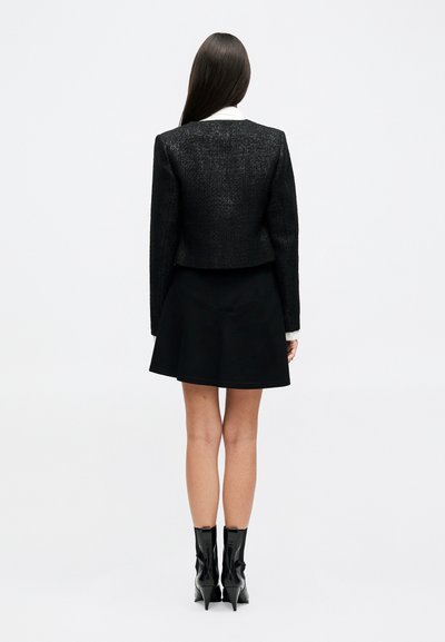 Žena s dlhými tmavými vlasmi stojí otočená chrbtom, má na sebe čiernu textúrovanú bundu, čiernu sukňu, biele manžety košele a čierne členkové topánky na opätku.