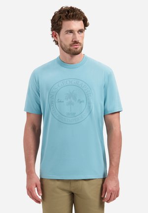 Jonge man met baard die een lichtblauw T-shirt met korte mouwen draagt met een subtiele palmboomprint en beige broek, kijkt naar rechts.