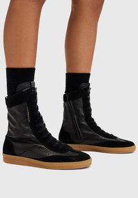 Sneakers alta in pelle nera e suede con dettagli perforati, suola in gomma e zip laterale. Presentano lacci neri spessi e polsino alla caviglia.