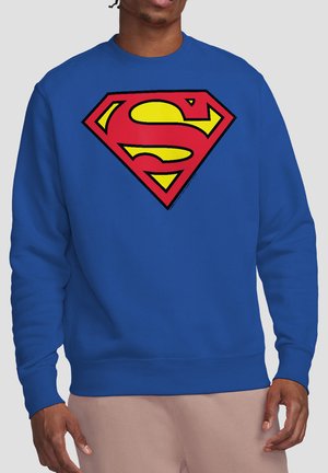 Mann trägt einen blauen Pullover mit einem großen roten und gelben Superman-Emblem auf der Brust und beigen Hosen.