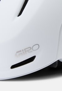 Weißer Fahrradhelm mit Belüftungsöffnungen und einem metallischen Logo an der Seite. Besitzt eine glatte, glänzende Oberfläche und eine abgerundete Form.