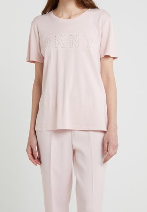 Kobieta ubrana w jasnoróżową koszulkę z krótkim rękawem z napisem "DKNY" ułożonym z małych białych koralików na piersi, w połączeniu z pasującymi spodniami.