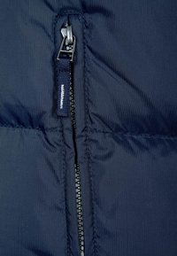 Marinblå puffjacka med tyg som har en framträdande dragkedja med en strukturerad, ribbad yta. Etikett fäst nära dragkedjan.