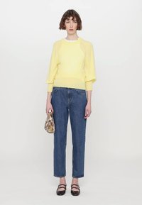 Gelber Strickpullover mit gepufften Ärmeln, kombiniert mit hochgemusterten blauen Jeans. Das Model hält eine gemusterte braune Handtasche.