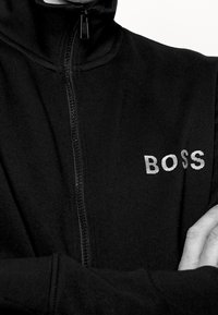Musta vetoketjullinen takki, jossa on korkea kaulus, teksturoitu kangas ja heijastava "BOSS"-logo vasemmassa rinnassa.