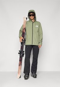 Olivgrüne, wasserdichte Jacke mit Kapuze, schwarze Hose und schwarze Skischuhe. Halte braune Skier mit Bindung in der Hand und stehe vor einem einfarbigen Hintergrund.