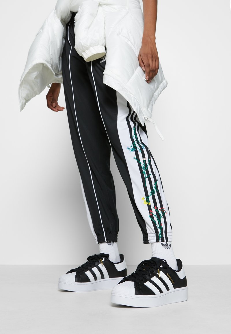 Mustat ja valkoiset verkkohousut, joissa on kukkakuvio sivussa, joustavat hihnat, yhdistetty mustiin ja valkoisiin lenkkareihin, joissa on kuorikärki ja kolme raitaa.