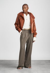 Chaqueta de cuero marrón con amplio cuello, suéter de canalé crema y pantalones gris con pliegues. El modelo lleva sandalias beige, mostrando un ajuste relajado.