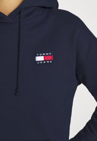 Sweat à capuche bleu marine avec cordons, arborant un petit patch logo rectangulaire Tommy Jeans en blanc, rouge et bleu marine sur la poitrine.