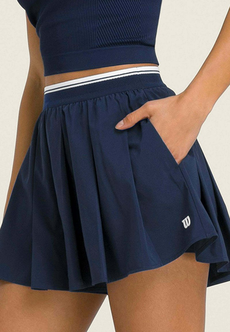 Femme portant une jupe plissée bleu marine avec la main dans la poche, associée à un top court assorti.