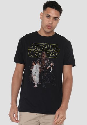 Junger Mann trägt ein schwarzes Star Wars T-Shirt mit den Figuren Luke Skywalker, Prinzessin Leia, Chewbacca und Han Solo auf der Vorderseite.