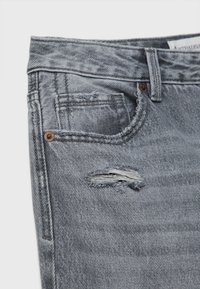 Jean en denim gris avec une déchirure horizontale effilochée près de la poche avant et des coutures visibles ainsi que des rivets métalliques.