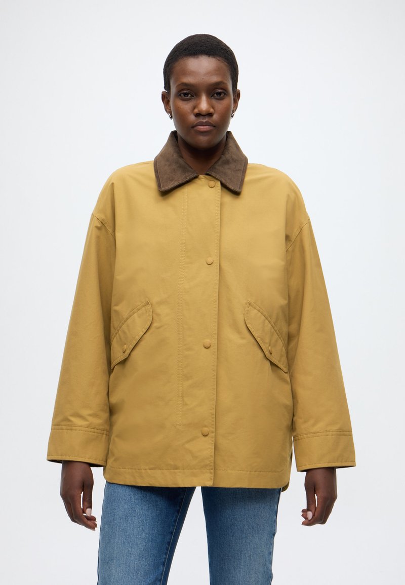 Veste extérieure jaune avec un col en daim marron, dotée d'une fermeture à boutons sur le devant et de deux poches latérales. Le tissu a une texture lisse.