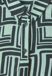 Koszula z kołnierzykiem w miętowym kolorze z czarnym abstrakcyjnym wzorem geometrycznym, z ciemnymi guzikami wzdłuż pliski oraz teksturowanym materiałem.