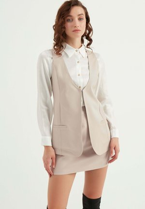 Beige vest over een witte blouse, met een v-hals, open voorkant en twee zakken, gecombineerd met een beige mini rok en zwarte kniehoog boots.
