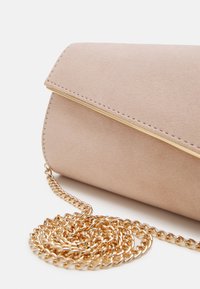 Pochette en daim beige avec bordure dorée et une bandoulière en chaîne dorée enroulée sur fond blanc.
