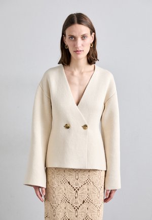 Cream-colored, textured jacket with a deep V-neck and bell sleeves. Features two gold buttons at the front. Worn over a lace skirt.