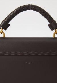 Sac en cuir marron foncé avec une poignée en cuir tressé et des accents en métal doré. Présente une marque en relief et des détails de couture visibles.