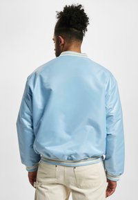 Blouson aviateur bleu clair avec une texture brillante, des poignets côtelés et un col rayé. Présente une coupe décontractée et un ourlet élastique.