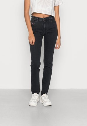 Personne portant un jean skinny noir taille haute, un top court blanc et des baskets blanches épaisses, debout devant un fond uni.
