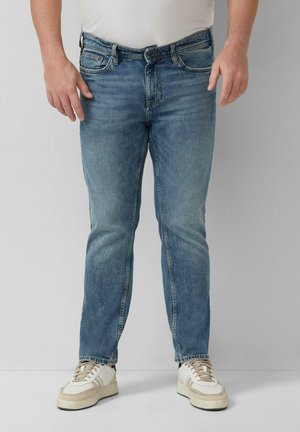 Mężczyzna w niebieskich dżinsach i białych trampkach stoi na tle gładkiej ściany, z rękami swobodnie opuszczonymi wzdłuż ciała.