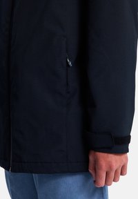 Schwarze Jacke mit einem Reißverschluss und einem Klettverschlussdetail an den Bündchen, hergestellt aus einem glatten Stoff mit matter Textur, dargestellt am Arm einer Person.