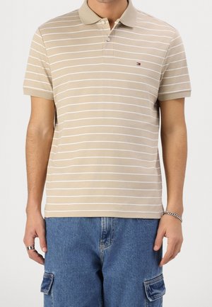 Polo beige à manches courtes avec fines rayures horizontales blanches porté par une personne portant également un jean cargo bleu et des bijoux aux mains.