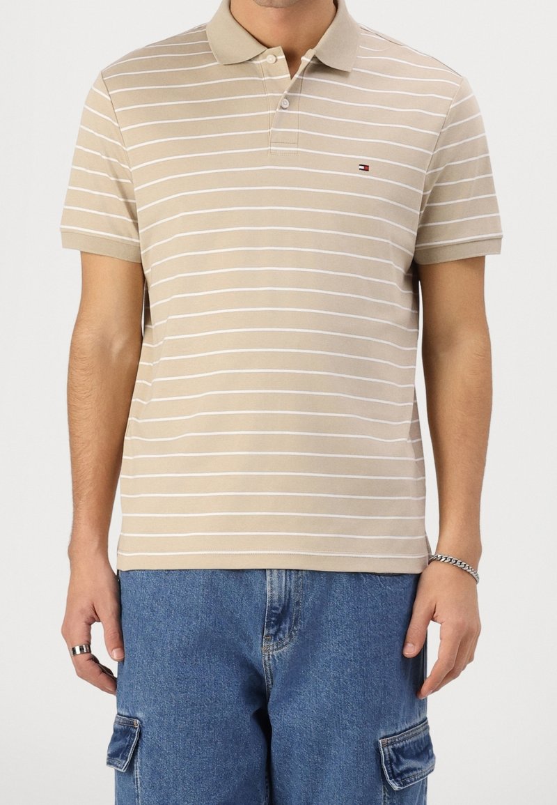 Polo beige à manches courtes avec fines rayures horizontales blanches porté par une personne portant également un jean cargo bleu et des bijoux aux mains.