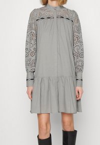 Vestido gris con mangas largas de encaje de ojales, acentuado por un dobladillo con volantes y bordados decorativos en los puños. Cierre frontal con botones.