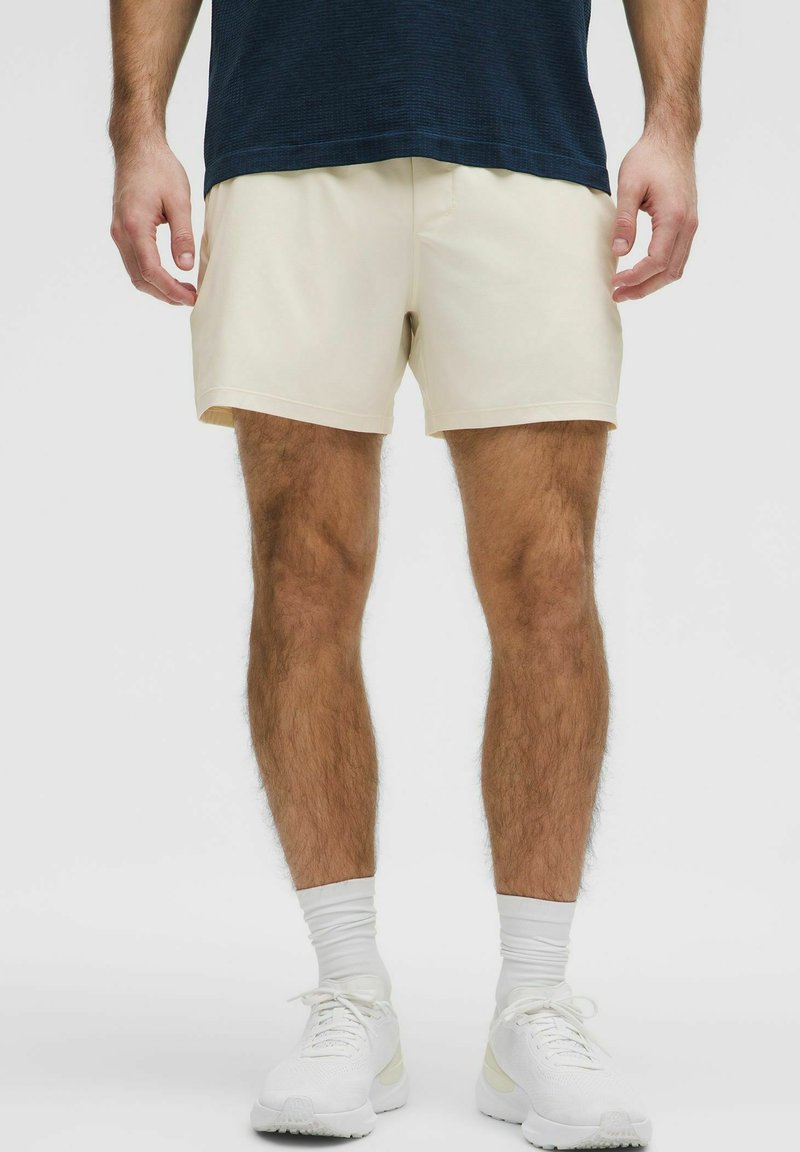 Cremefarvede shorts med en glat tekstur, elastisk talje og skræddersyet pasform, parret med hvide træningssko og lyse sokker.