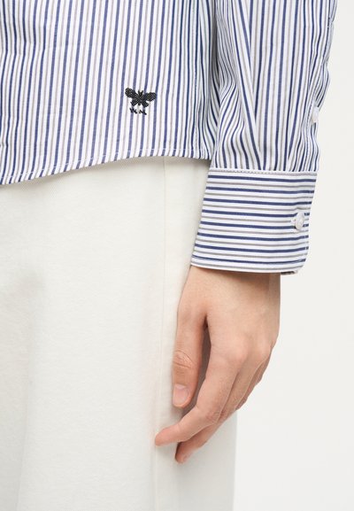 Χέρι που ακουμπάει στο πλάι, φορώντας μπλε και λευκό ριγέ πουκάμισο με μια μικρή μαύρη κεντημένη μέλισσα πάνω από κρεμ παντελόνι.