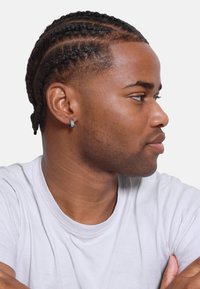 Perfil de una persona con cabello trenzado que lleva un arete de aro plateado. Está vestida con una camiseta blanca sencilla frente a un fondo blanco sólido.
