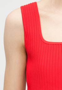 Primer plano del hombro y la parte superior del pecho de una persona que lleva una camiseta sin mangas de color rojo brillante con textura acanalada y escote cuadrado.