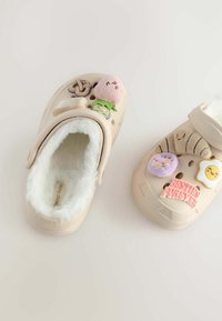 Sabots beiges en fourrure avec des patchs décoratifs comprenant des aliments et des phrases, dotés de trous de ventilation circulaires et d'un design confortable.