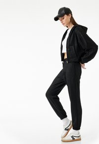 Sudadera corta negra brillante con cremallera frontal, combinada con pantalones jogger negros, rematada con zapatillas blancas con acentos negros.
