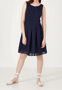 Námorně modré šaty bez rukávů s sklady, délka po kolena a textilní materiál. Kombinováno s bílými espadrilkami s kotníkovými vázáními.