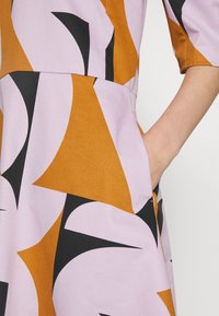 Gros plan sur une robe à motif géométrique en moutarde, lavande et noir avec une main placée dans la poche latérale.