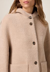 Abrigo de mezcla de lana beige con un acabado texturizado, capucha y tres grandes botones marrones; cuenta con bolsillos laterales para mayor funcionalidad.