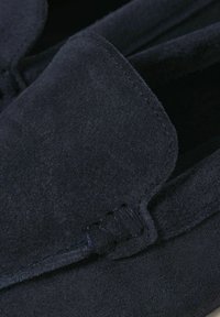 Prim-plan al unei mocasine negre din piele intoarsa, evidentiind cusaturile detaliate si partea superioara de tip mocasin, realizata din piele texturata si moale.