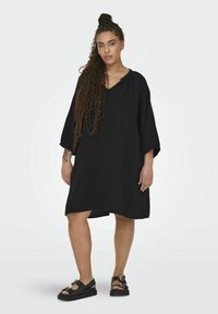 Black, knee-length dress made of textured fabric with a loose fit, wide sleeves, and a V-neckline. Worn with black platform sandals.