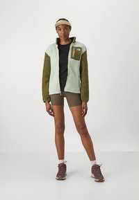 Fleece jacket in light green and olive green, features zip front, side pockets, and a zippered chest pocket; paired with black top and shorts.