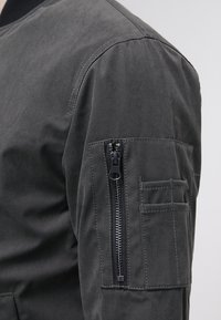 Giubbotto bomber grigio scuro con un tessuto testurizzato, colletto a costine e una tasca con zip sulla manica. Include un ulteriore taschino con patta.