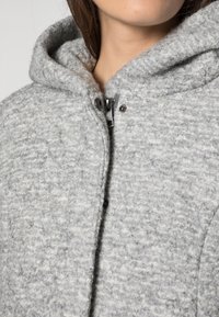 Siva kapuca jakna iz teksturirane tkanine, s prednjim patentnim zatvaračem od metalne opreme in mehko notranjostjo.