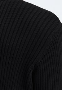 Gros plan sur un tissu tricoté côtelé noir montrant des lignes verticales texturées et des coutures détaillées sur l'épaule et la manche d'un vêtement.